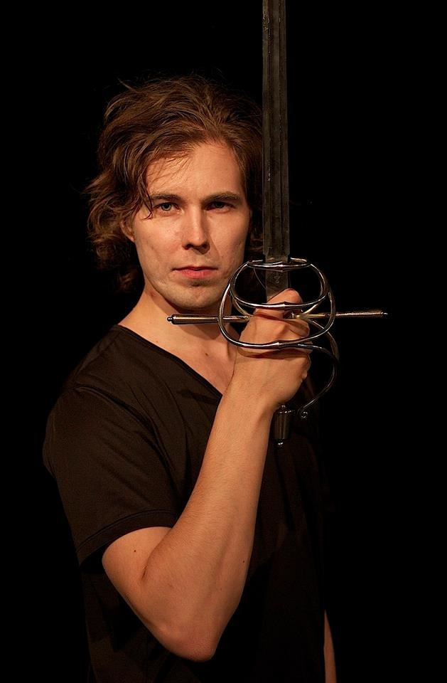 Juuso-Matias with a musketeer sword held vertically. Dark background, black t-shirt and brown hair.