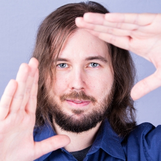 Juuso-Matias forming a triangle with their hands. Violet, blue shirt and brown hair, beard and piercing-grey eyes.